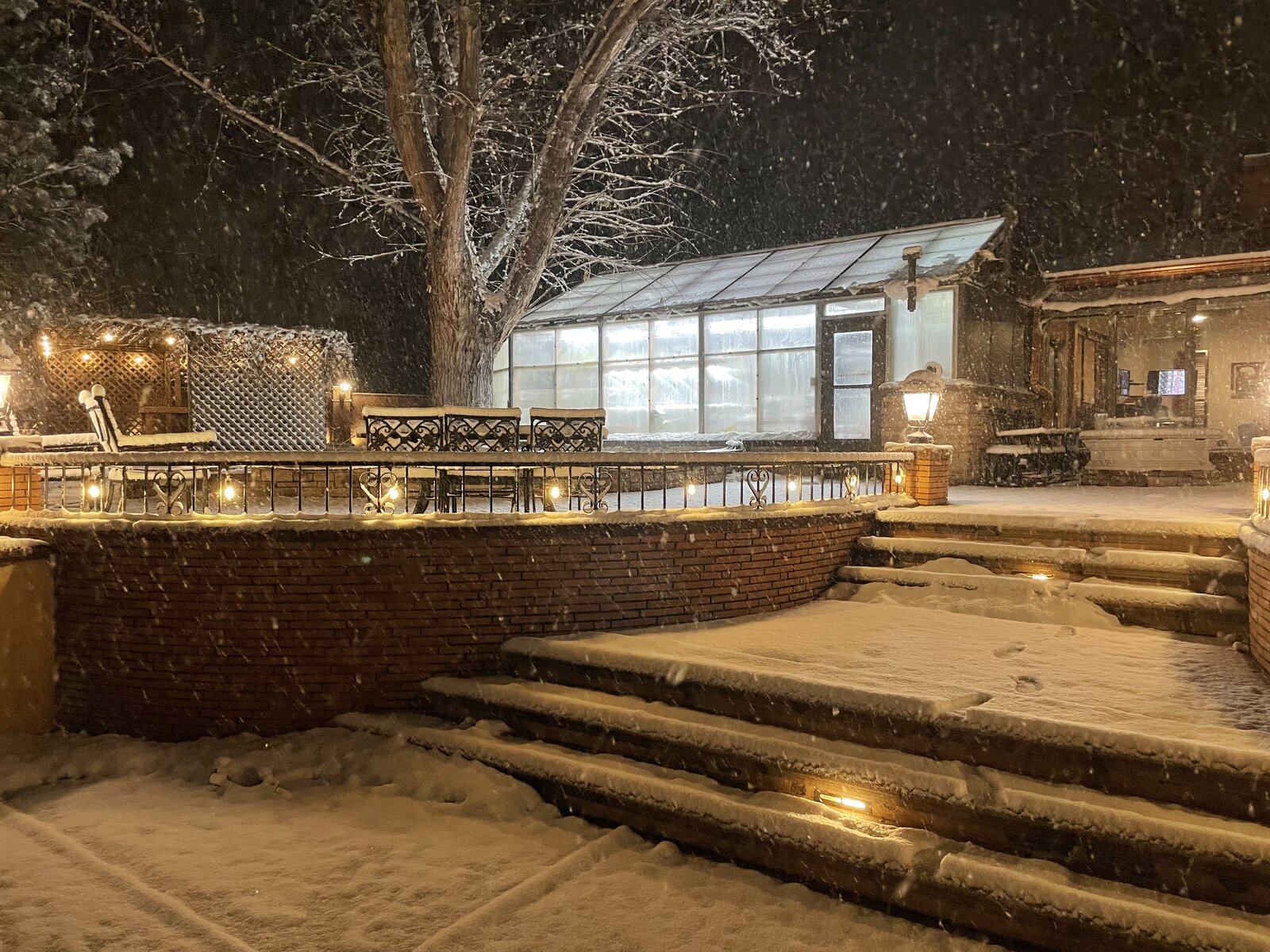 Greenhouse exterior at night during snowfall — the hexagonal shape, polycarbonate panels, and brick base are visible
